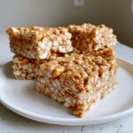 Four squares of chewy peanut butter rice krispie treats stacked on a white plate.