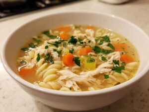A close-up of a white bowl filled with hot penicillin soup featuring shredded chicken, egg noodles, carrots, and parsley.