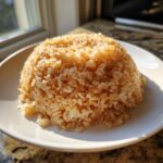 A perfectly molded mound of seasoned sushi rice, glistening under sunlight, served on a white plate.