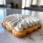 Close-up of a golden sugar cookie topped with thick, white, piped sugar cookie icing and tiny sprinkles.