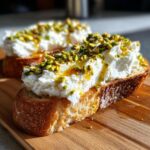 A close-up of delicious ricotta toast topped with whipped ricotta, chopped pistachios, and a drizzle of honey.