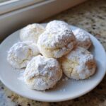 A plate of soft, round cool whip cookies heavily dusted with powdered sugar, sitting on a granite countertop.