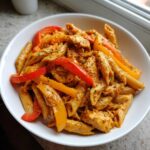 A white bowl filled with creamy, orange-hued Rasta pasta mixed with chicken pieces and colorful red and yellow bell peppers.