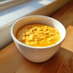 A close-up of a vibrant orange beer cheese dip, sprinkled with black pepper, served in a white bowl.