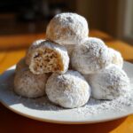A mound of freshly baked russian tea cakes heavily coated in powdered sugar, one cookie is broken open showing the crumb.
