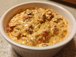 Close-up of a rich, orange-hued sausage cheese dip mixed with tomatoes and peppers in a white bowl.