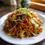 A close-up of a generous serving of savory fried noodles mixed with cabbage, carrots, and mushrooms, topped with scallions and sesame seeds.