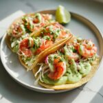Three flavorful shrimp taco servings piled high with pink shrimp, cabbage slaw, and drizzled with creamy green sauce.