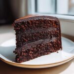 A decadent slice of three-layer chocolate fudge cake covered in smooth, dark ganache frosting, sitting on a white plate.