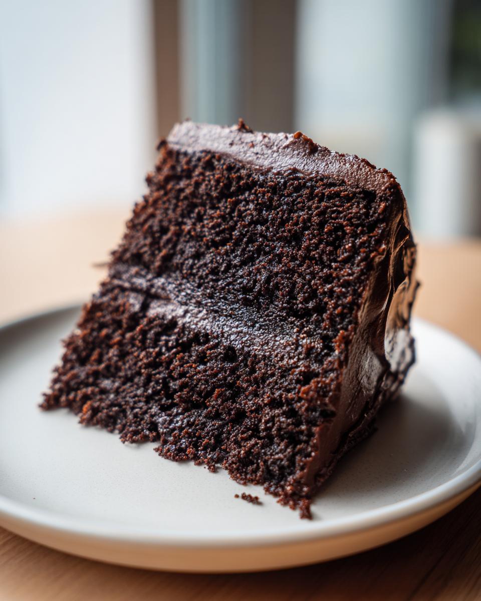 A close-up of a moist, dark slice of chocolate fudge cake with thick frosting on a light plate.