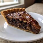 A close-up shot of a decadent slice of chocolate pecan pie resting on a white plate.