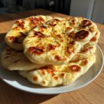 A stack of four freshly made, soft naan bread pieces, brushed with melted butter, garlic, and herbs, sitting on a white plate.