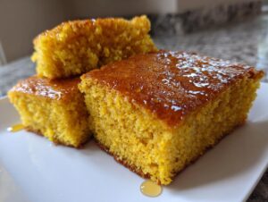 Three golden yellow slices of moist sourdough cornbread drizzled generously with honey on a white plate.