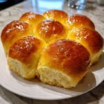 A cluster of freshly baked, golden brown sourdough discard rolls with a shiny glaze, arranged on a white plate.