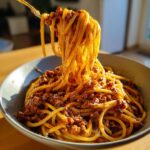 A fork lifting long strands of spaghetti coated generously with rich bolognese sauce from a grey bowl.