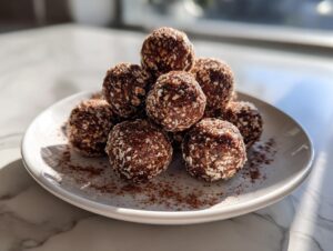 A stack of homemade chocolate protein balls coated in oats, dusted with cocoa powder on a white plate.