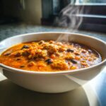 Close-up of a steaming white bowl filled with rich, orange pumpkin chili featuring ground meat and black beans.