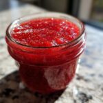 Close-up of bright red strawberry freezer jam filling a small glass jar, showing visible seeds.