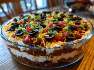 A close-up of a stunning 7 layer dip, topped with shredded cheese, tomatoes, green onions, and black olives.