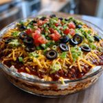 Close-up of a stunning 7 layer dip in a glass bowl, topped with cheese, olives, tomatoes, and green onions.