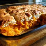 A close-up view of a freshly baked sweet potato cobbler showing the bright orange filling and golden biscuit topping.