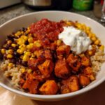 A close-up of a sweet potato taco bowl featuring roasted sweet potato cubes, brown rice, black beans, corn, salsa, and sour cream.