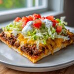 A close-up slice of taco pizza featuring seasoned beef, melted cheese, shredded lettuce, diced tomatoes, and sour cream.