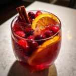 A glass filled with a vibrant red thanksgiving punch, garnished with fresh cranberries, orange slices, and cinnamon sticks.