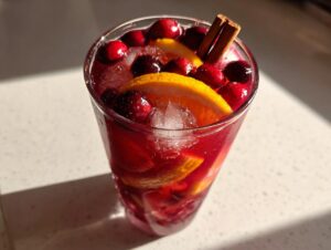 Close-up of a glass filled with vibrant red thanksgiving punch, ice, orange slices, cranberries, and cinnamon sticks.