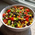 A close-up of a white bowl filled with colorful cowboy caviar salad featuring black-eyed peas, corn, tomatoes, and peppers.