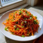 A bright orange mound of shredded raw carrot salad topped with sesame seeds and chopped green onions.