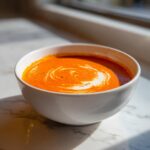 A vibrant orange bowl of tomato bisque topped with a swirl of cream, sitting on a marble surface.