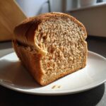 Cross-section of a rustic whole wheat bread loaf showing a soft crumb texture and seeds, resting on a white plate.