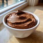 Close-up of a thick, rich chocolate dip swirled high in a small white bowl, set near a sunlit window.
