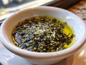Close-up of a white bowl filled with flavorful olive oil bread dip, featuring minced garlic and green herbs.