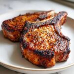 Close-up of two beautifully seared air fryer pork chops with a dark, caramelized crust served on a light plate.