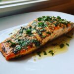 A close-up of a glistening, cooked fillet of air fryer salmon topped with minced garlic and fresh parsley, resting in butter sauce.