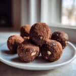 A stack of rich, dark Almond Joy Truffles generously dusted with cocoa powder, resting on a white plate.