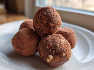 A stack of four rich, dark chocolate Almond Joy truffles dusted heavily with cocoa powder on a white plate.