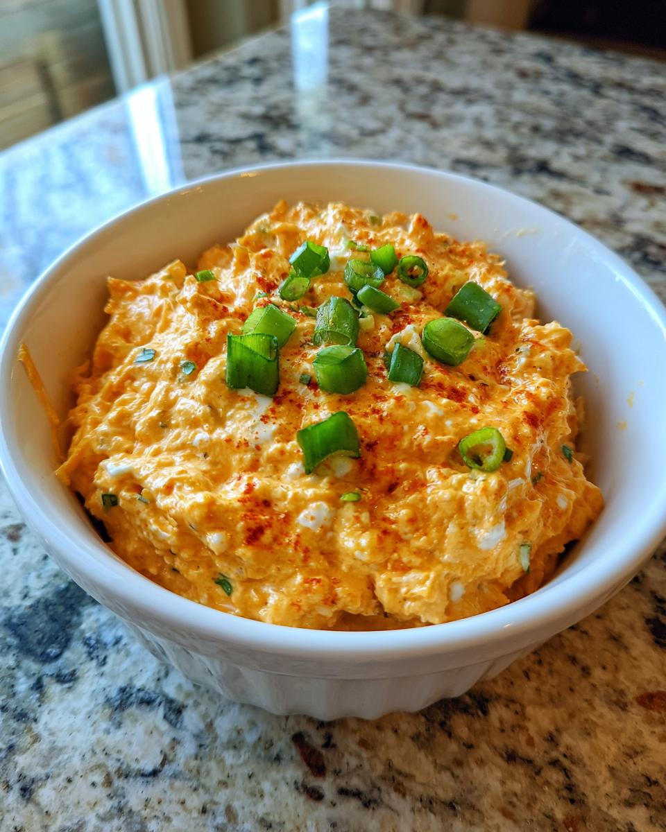 A close-up of creamy, orange-colored boat dip topped with chopped green onions and paprika in a white bowl.