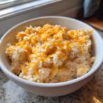 A white bowl filled with creamy cheesy rice topped with freshly shredded, slightly melted cheddar cheese.