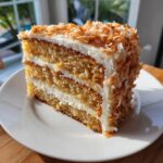 A close-up of a moist, three-layer coconut cake slice covered in white frosting and toasted coconut flakes.