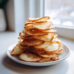 A tall stack of golden brown, homemade potato chips sprinkled with salt on a white plate.