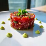 A beautifully molded stack of vibrant red tuna tartare garnished with herbs and surrounded by dots of green avocado cream.