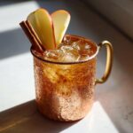 A frosty copper mug filled with an apple cider Moscow mule cocktail, garnished with apple slices and cinnamon sticks.