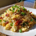 A serving of savory cauliflower rice mixed with scrambled egg and bacon pieces, topped with green onions.