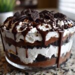 Close-up of a layered Baileys Chocolate Cheesecake Trifle topped with whipped cream, chocolate sauce, and crushed cookies.