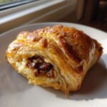 A golden, flaky piece of baked brie in puff pastry, showing melted cheese and a sweet pecan topping.
