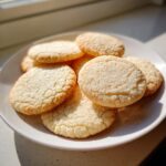 A close-up of several perfectly baked vegan sugar cookies stacked on a white plate, highlighted by bright sunlight.