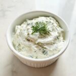 Close-up of creamy homemade tartar sauce, speckled with green herbs and topped with a sprig of fresh dill.